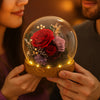 Two people holding a glass dome with red and purple roses inside, against a warm background.