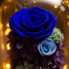 Blue rose and other preserved flowers in a decorative glass holder with a warm glow.