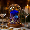 Decorative arrangement of blue and purple flowers under a glass dome on a table setting with wine glasses and candles.