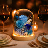 Decorative glass dome with blue and white flowers on a table setting with wine glasses and plates.
