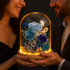 Two people holding a glass dome with blue roses and flowers inside, illuminated from below.