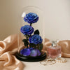 Three blue roses under a glass dome with a perfume bottle and pearls on a beige fabric background