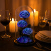 Blue roses under a glass dome on a romantic dinner table with candles and wine glasses.