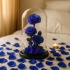 Three blue roses under a glass dome on a bed with scattered rose petals and a lit candle.