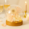 Glass dome with preserved flowers on a wooden base, set against a blurred background with glasses and candles.
