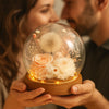 Two people holding a glass dome with preserved flowers and lights inside.