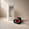 White gift box with a clear window next to a red preserved rose arrangement on a beige surface.