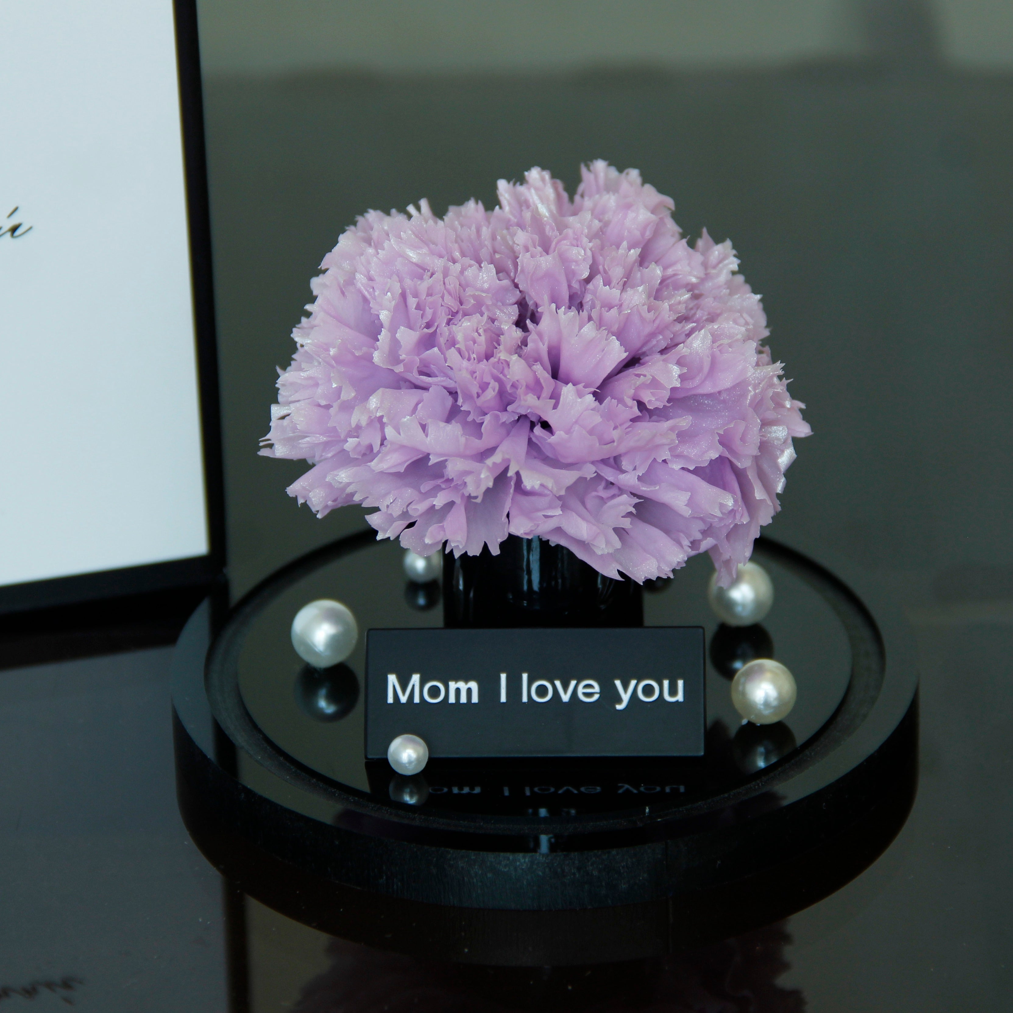 Purple flower arrangement on a black base with 'Mom I love you' card on a reflective surface.
