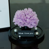 Purple flower arrangement on a black base with 'Mom I love you' card on a reflective surface.