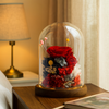 Glass dome with preserved flowers on a wooden table next to a lamp.