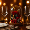 Decorative arrangement of preserved flowers under a glass dome on a table with wine glasses and plates.