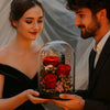 Man and woman holding a glass dome with red preserved roses and small flowers.