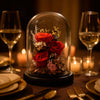 Decorative glass dome with red preserved roses on a table setting with wine glasses and candles.
