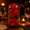Red preserved roses under a glass dome on a romantic dinner setting with candles and wine glasses.