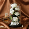 Glass dome with white preserved roses, perfume bottle, and necklace on a brown fabric background